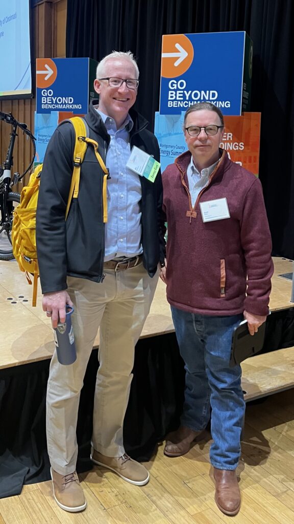 Two conference attendees standing together in front of ‘Go Beyond Benchmarking’ signage, wearing name badges and smiling at an energy industry event.